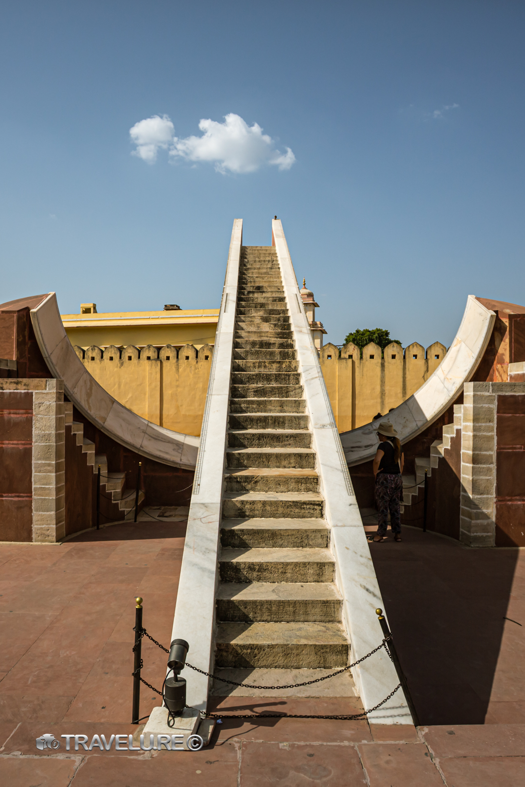 Smaller yantra instrument at Jantar Mantar Jaipur, symmetrical gnomon staircase pointing skyward with single cloud above, visitor at right for scale, Rajasthan, India.